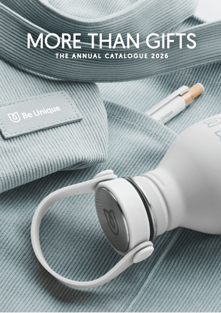 Catalog cover showing a Be Unique tag on blue knit fabric, white over-ear headphones, and a capped product; text reads 'MORE THAN GIFTS' and 'THE ANNUAL CATALOGUE 2026'.